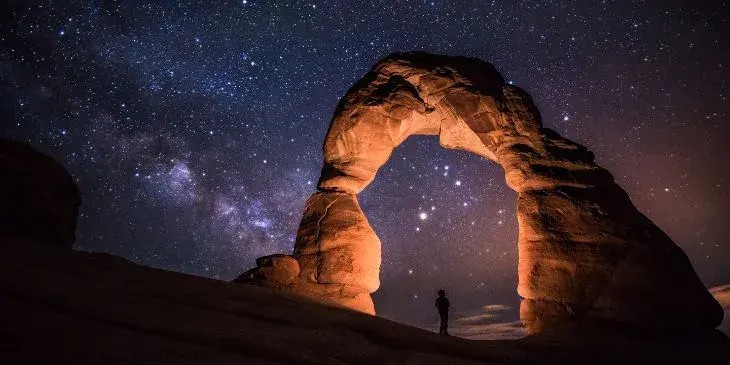 Uma pedra em formato de portal com um ceu estrelado a noite com uma pessoa no meio da pedra