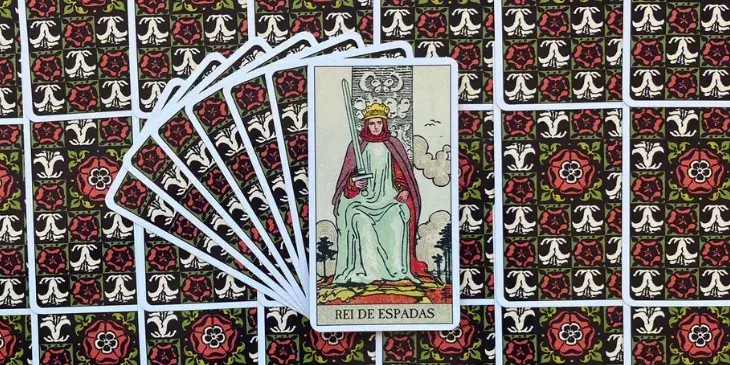 Rei está retratado sentado em um trono, erguido no alto sob um céu cinzento e tumultuoso, o que pode representar os desafios e as tempestades que ele enfrenta e supera com sua mente aguçada. Ele segura uma espada apontada para cima em uma mão, um símbolo clássico de poder e decisão, indicando sua capacidade de usar a lógica e o julgamento para cortar através da confusão e alcançar a verdade. A espada também sugere a comunicação clara e objetiva, características essenciais de um líder eficaz