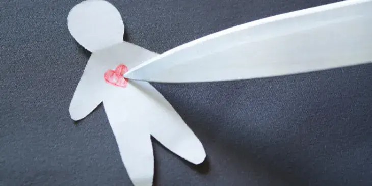 um homem feito de papel branco com um cora��o vermelho desenhado no peito com uma faca encostada.