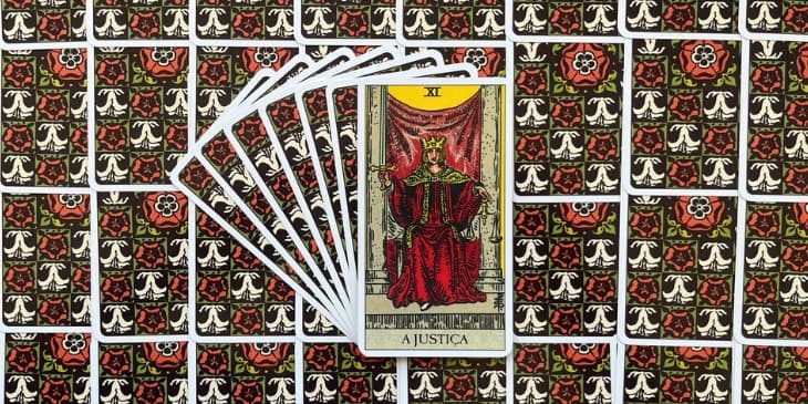 uma pessoa sentada em um trono ao lado de dois pilares, usando um manto vermelho e uma coroa de ouo, na m�o direita segura uma espada apontada para o ceu e na m�o direita segura uma balan�a.