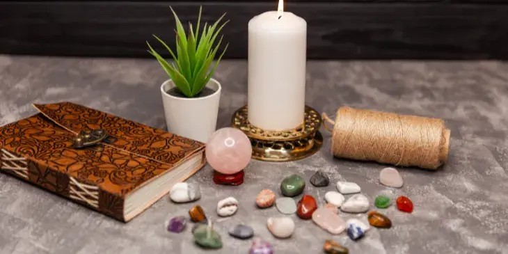 uma mesa com uma vela branca, um vaso de flor, um livro de capa marrom e varias pedrinhas coloridas.