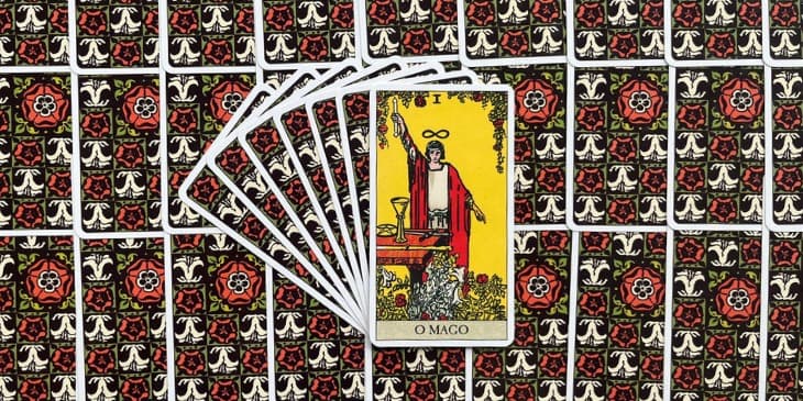carta do mago com uma mao apontada para o ceu e a outra para a terra com seu manto vermelho e na mesa de madeira os elementos das cartas de tarot, o c�lice, bast�o espadas e ouros.