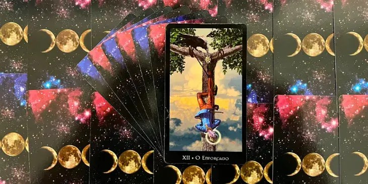 Tarot das Bruxas nos traz a imagem de um jovem sereno, suspenso de cabe�a para baixo por um p� em um freixo, uma �rvore forte e resistente, O rosto sereno e tranquilo do jovem contrasta com sua posi��o invertida, sugerindo que, apesar do desconforto e da desorienta��o que essa posi��o pode inicialmente causar, h� um processo de aceita��o e compreens�o em andamento. Esse contraste � refor�ado pelo halo que rodeia a cabe�a do jovem, indicando a epifania ou a revela��o que est� experimentando. A perspectiva invertida se torna, assim, um instrumento de conhecimento e autoconhecimento. Ao fundo, um c�u tranquilo e apraz�vel transmite a mensagem de que, apesar da aparente dificuldade da situa��o, n�o h� motivo para temer. Esta fase de transi��o � necess�ria e ben�fica, mesmo que seja desconfort�vel.