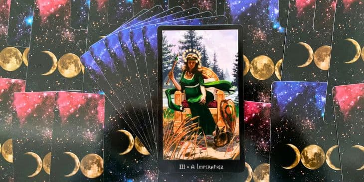 carta do tarot das bruxas a imperatriz onde mostrar a figura de uma mulher sentada em um trono de vestido verde e uma coroa, ao lado do trono tem um escudo e um coelho ao ar livre
