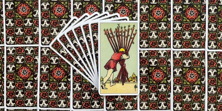 um homem andando de costas e carregando 10 bastoes de paus com muita dificuldade, atrás tem uma pequena cidade medieval.