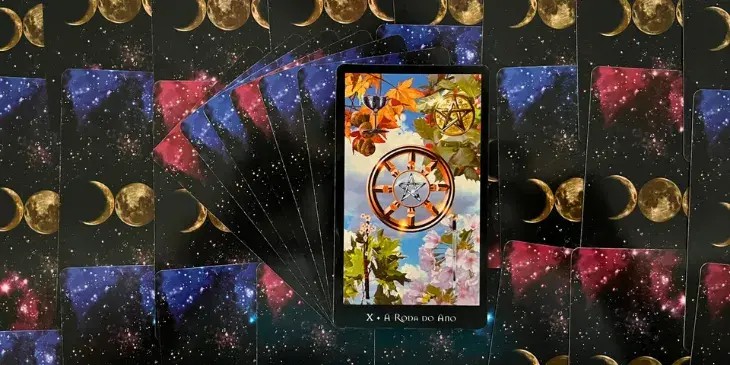 No centro da carta, um pentagrama lunar e prateado - um emblema ancestral do poder feminino e do mist�rio da lua - � emoldurado pela Roda do Ano dourada e solar, simbolizando a passagem do tempo, os ciclos da vida e o equil�brio entre luz e escurid�o. O dourado � associado ao masculino, ao sol e a tudo que � consciente e manifesto. Ao redor da Roda do Ano, oito raios representam os oito sabbats - dias sagrados para as bruxas, que marcam momentos significativos nos ciclos naturais e sazonais. No fundo, o c�u azul e as nuvens suaves evocam a vastid�o do universo e a leveza do esp�rito, criando uma atmosfera de harmonia e tranquilidade. As quatro esta��es do ano e seus correspondentes naipes do tar� se apresentam no entorno da Roda. Cada um deles est� ligado a um dos quatro elementos (ar, terra, fogo e �gua), representando diferentes aspectos da experi�ncia humana e do cosmos. No topo da Roda, a simbologia do elemento terra e o Solst�cio de Inverno s�o representados pela moeda e pelo acebo, uma planta associada � Natividade e frequentemente usada para prote��o e boa sorte. Essa combina��o invoca uma sensa��o de estabilidade, for�a e perseveran�a diante do frio e das adversidades. Na parte inferior direita, o Equin�cio da Primavera � representado por flores rosadas de cerejeira, que se aliam � espada, s�mbolo do elemento ar. Este conjunto evoca a renova��o da vida, a for�a do pensamento e a nobreza de car�ter, expressando a transi��o da introspec��o do inverno para a expans�o da primavera. Na parte inferior esquerda, vemos o Solst�cio de Ver�o representado por folhas verdes de carvalho e uma vara de espinho em flor, representando o elemento fogo. Simbolizam a plenitude da vida, a sabedoria e a virtude, refor�ando o sentimento de crescimento e alegria que o ver�o traz. Por fim, na parte superior esquerda, as folhas alaranjadas de bordo e as bolotas da colheita representam o Outono e est�o alinhadas ao naipe de copas, associado ao elemento �gua. As folhas de bordo refletem a beleza e a energia da mudan�a, enquanto as bolotas sugerem prosperidade e sabedoria, representando a colheita ap�s um ciclo de trabalho e crescimento.
